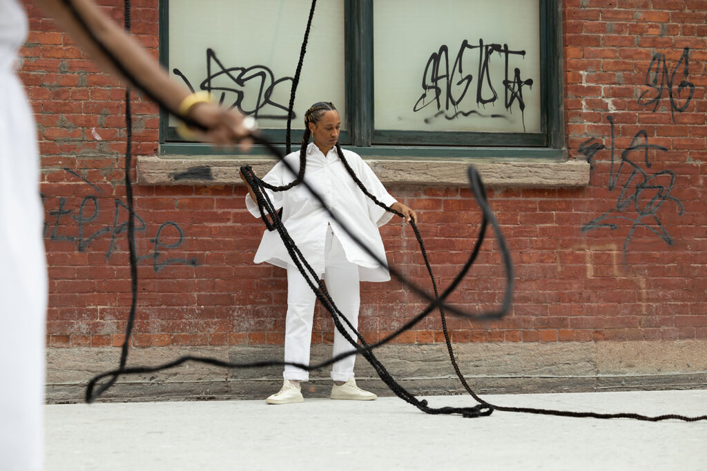 A person standing against a brick wall holding extended braids.