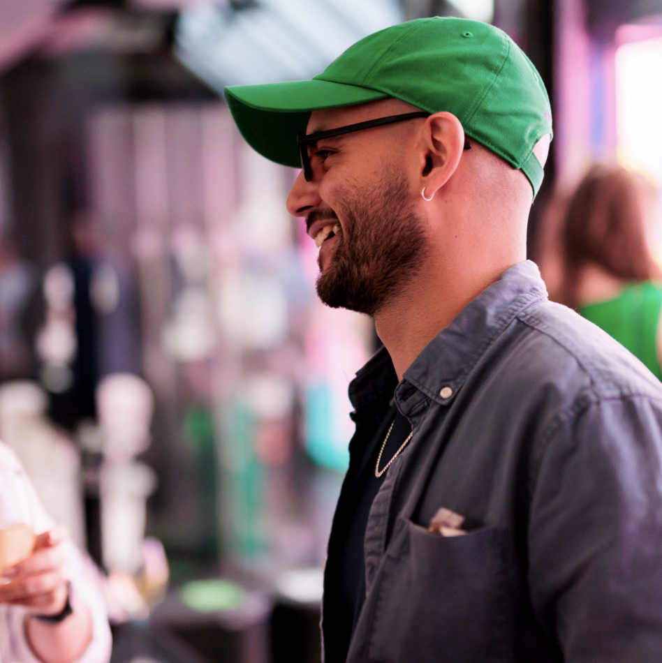 Side profle picture of a man wearing an green hat, black glasses and smiling.